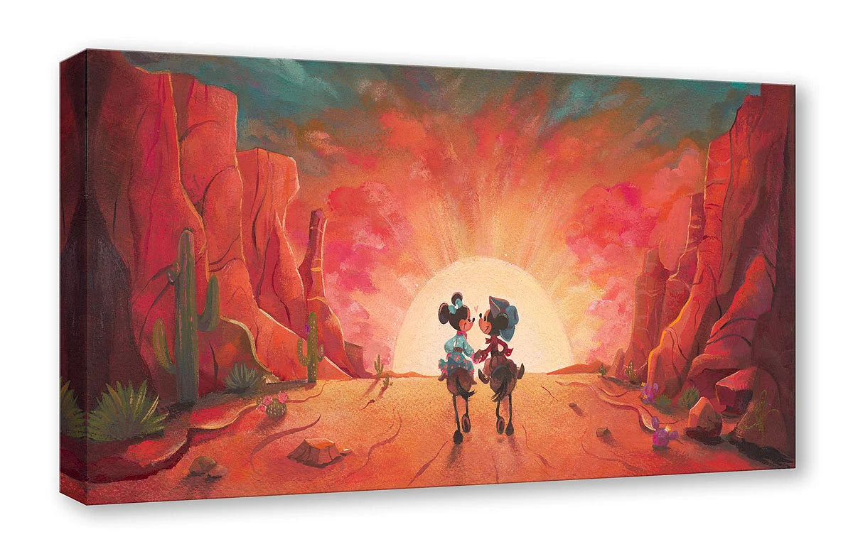 A rectangular canvas wall art print, shown at an angle, features a stylized desert canyon scene with two small figures riding toward a glowing sun between red rock cliffs.